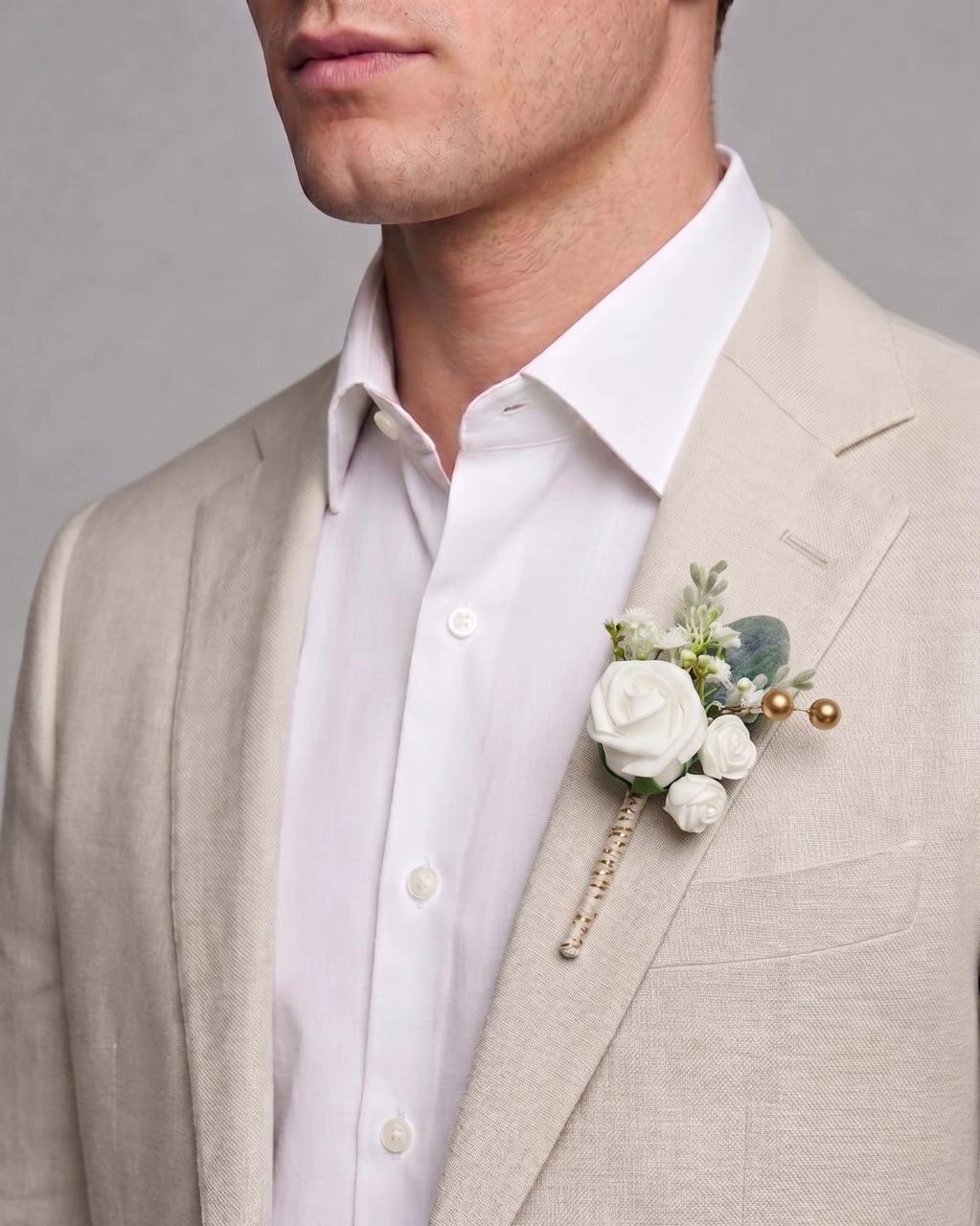 A close-up of an ivory pre-made boutonniere secured on a man's beige suit lapel.